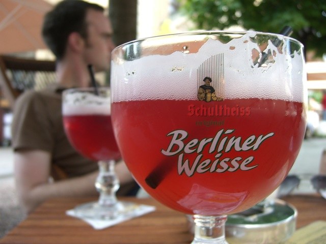 a glass of Schultheiss Berliner Weisse beer on a wooden table in a cafe, outdoors, against a blurred background of visitors